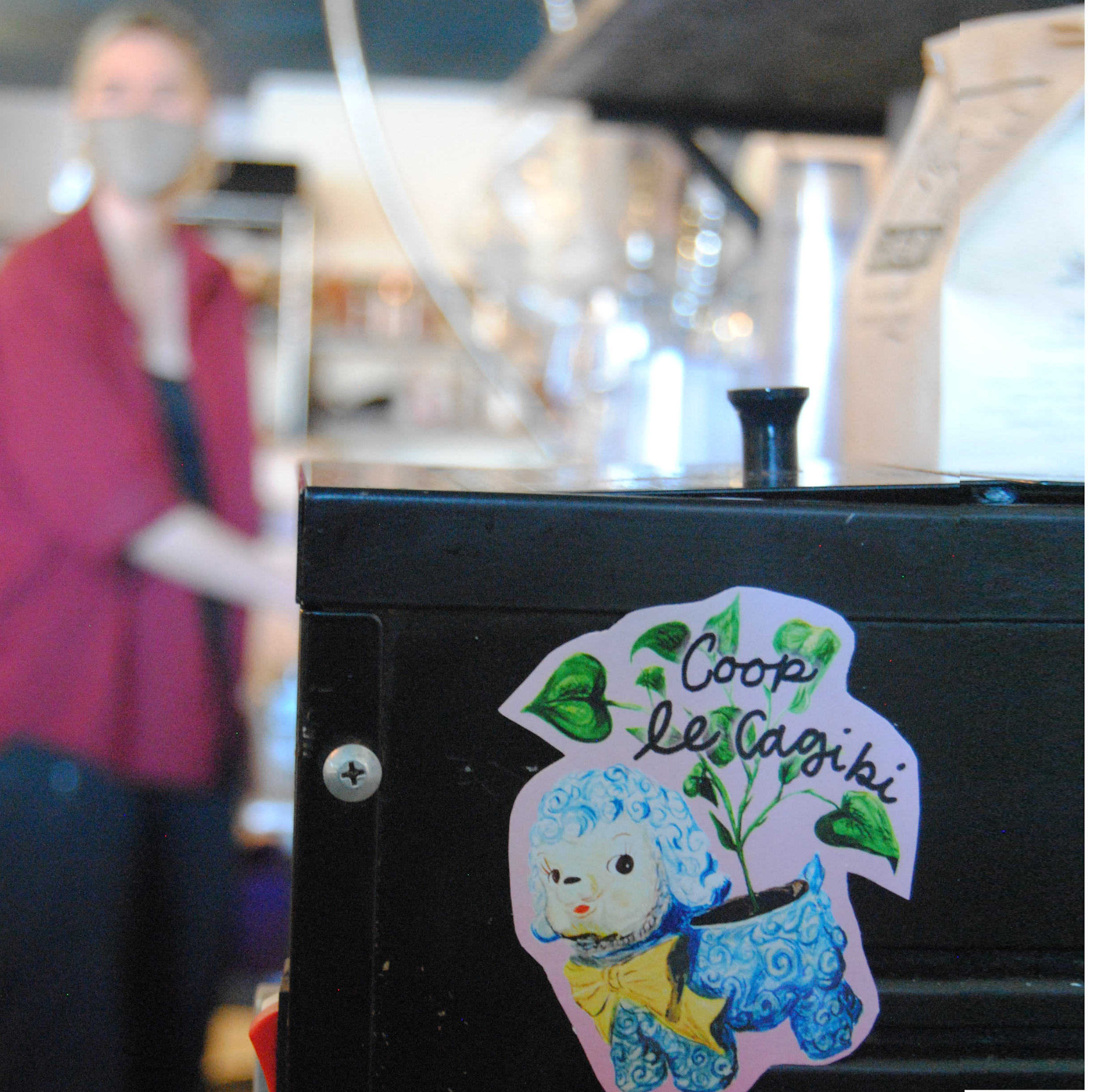 photo d'un-e barista à l'arrière plan et d'un autocollant du cagibi sur une machine a café au premier plan